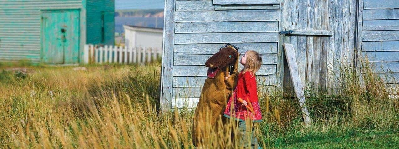 A child stands beside a person in a bear costume near a weathered wooden building with tall grass in the foreground. Rural landscape with more wooden structures and a white fence in the background.